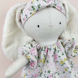 May include: A white bunny doll with long, fluffy ears, wearing a pink and green floral headband and a matching dress with a gathered neckline.