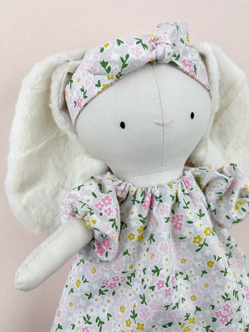May include: A white bunny doll with long, fluffy ears, wearing a pink and green floral headband and a matching dress with a gathered neckline.