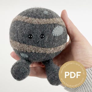 Op de afbeelding: Een handgemaakt grijs pluche planeetspeelgoed met een lachend gezicht, bruine strepen en kleine grijze vlekken. Het speelgoed heeft twee kleine poten en wordt in een hand gehouden. Een cirkelvormige sticker met de tekst "sewing pattern and tutorial PDF" staat in de rechter benedenhoek.