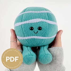 Op de afbeelding: Een zacht, turquoise speelgoed in de vorm van een planeet met witte strepen, zwarte ogen en een gestikt glimlachje. Het speelgoed heeft kleine turquoise benen en wordt in twee handen gehouden. Een cirkelvormige afbeelding met de tekst "sewing pattern and tutorial PDF" staat linksonder.
