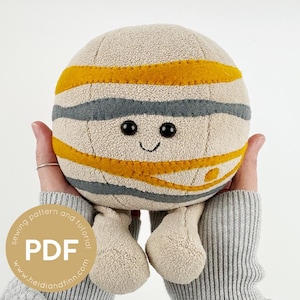 Op de afbeelding: Een zacht planetenspeelgoed met een lachend gezicht, zwarte ogen en beige, grijze en gele strepen. Het speelgoed wordt in twee handen gehouden. De tekst "PDF" is zichtbaar op een ronde sticker.