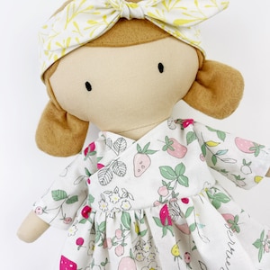 May include: A handmade doll with brown hair, wearing a white dress with a strawberry print and a yellow headband with a leaf pattern.
