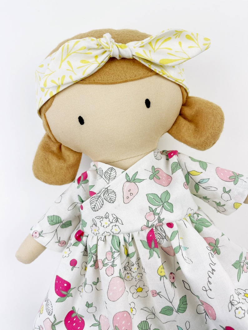 May include: A handmade doll with brown hair, wearing a white dress with a strawberry print and a yellow headband with a leaf pattern.