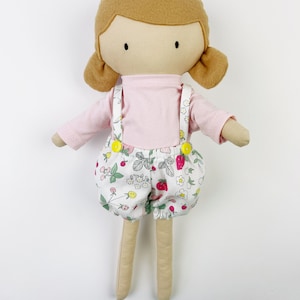 May include: A handmade doll with brown hair, a pink shirt, and white overalls with a strawberry print. The doll has a simple face with black eyes and a small smile.