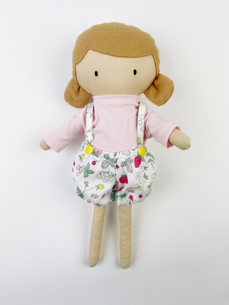 May include: A handmade doll with brown hair, a pink shirt, and white overalls with a strawberry print. The doll has a simple face with black eyes and a small smile.