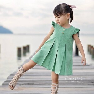 May include: A young girl wearing a green pleated dress with a button closure and ruffle sleeves. She is wearing beige gladiator sandals and is standing on a wooden dock.