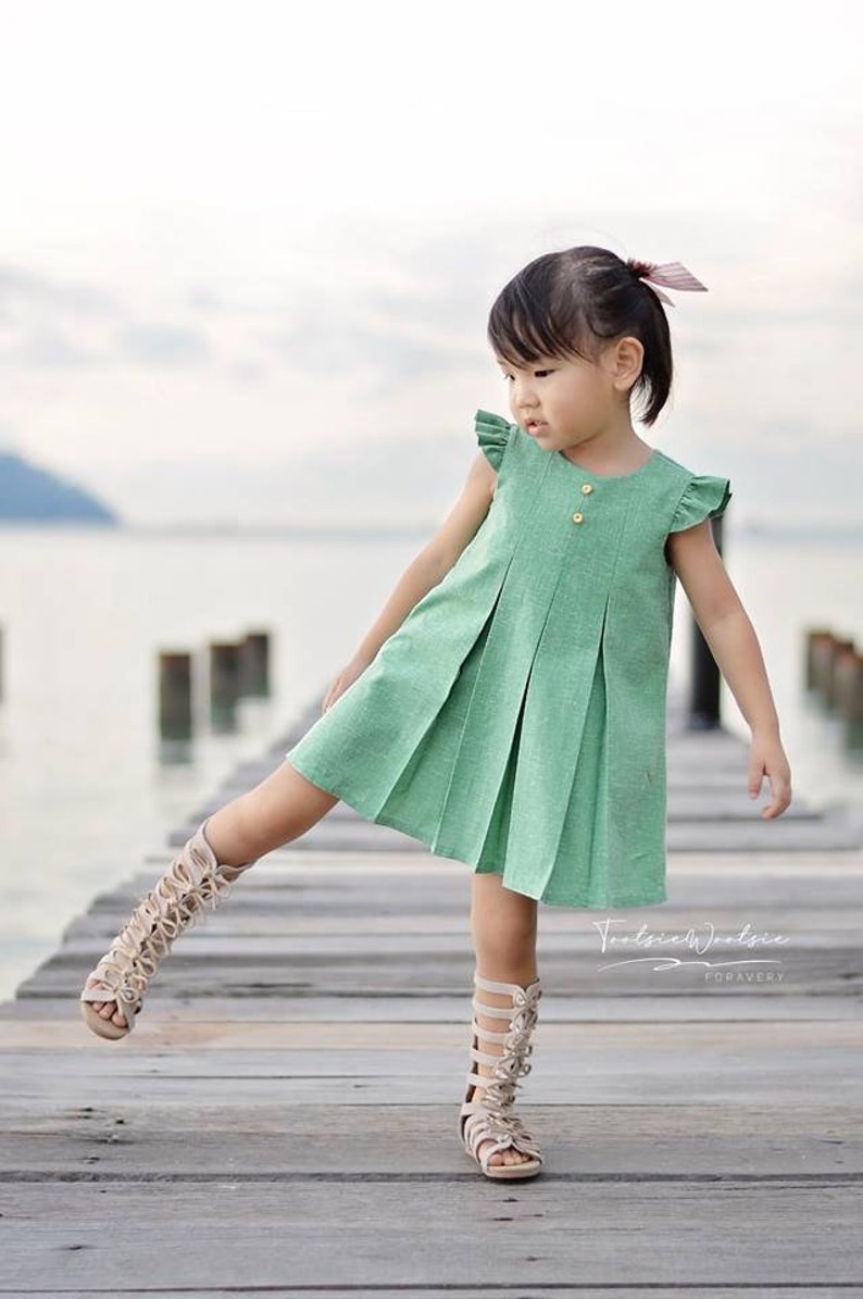 May include: A young girl wearing a green pleated dress with a button closure and ruffle sleeves. She is wearing beige gladiator sandals and is standing on a wooden dock.