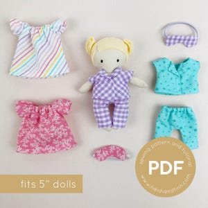May include: A doll in purple checked pyjamas, with additional doll clothing including a striped top, pink dress, blue star-patterned pyjamas, and a sleep mask. The image includes the text "fits 5" dolls".