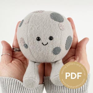 Könnte beinhalten: Ein graues Mond-Plüschtier mit einem lächelnden Gesicht und dunkelgrauen und hellrosa Flecken. Das Spielzeug hat zwei kleine graue Beine und wird in offenen Händen gehalten. Ein PDF-Schnittmuster und Tutorial sind verfügbar.