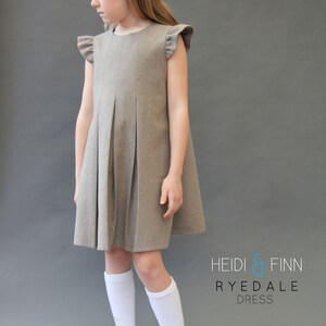 May include: A young girl wearing a brown pleated dress with ruffled sleeves and white knee-high socks. The dress has a simple design and is made of a soft, comfortable fabric. The text "HEIDI & FINN RYEDALE DRESS" is printed on the bottom right corner of the image.