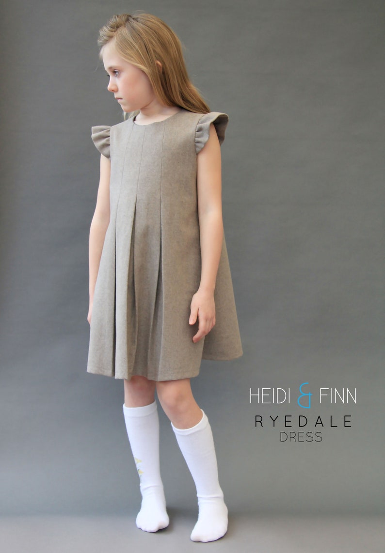 May include: A young girl wearing a brown pleated dress with ruffled sleeves and white knee-high socks. The dress has a simple design and is made of a soft, comfortable fabric. The text "HEIDI & FINN RYEDALE DRESS" is printed on the bottom right corner of the image.
