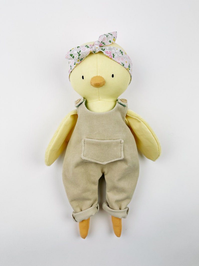 May include: A yellow plush chick toy wearing a floral headband and beige overalls. The chick has a yellow beak and black eyes.
