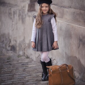 May include: A young girl wearing a black beret, a gray plaid dress, white tights, and black boots stands next to a brown leather satchel.