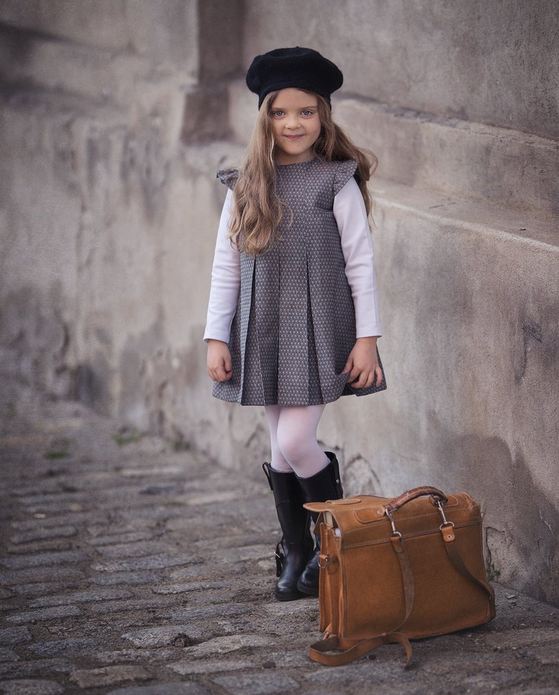 May include: A young girl wearing a black beret, a gray plaid dress, white tights, and black boots stands next to a brown leather satchel.