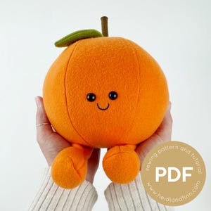 May include: A soft toy orange with a smiling face, green leaf, and brown stem. The orange toy has two small orange feet. The toy is held in a person's hands. The image includes a PDF sewing pattern and tutorial.