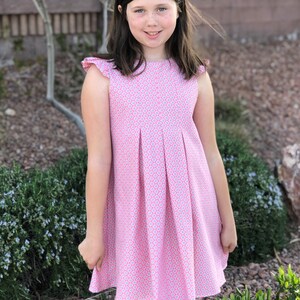 May include: A young girl wearing a pink and white patterned dress with a ruffled neckline. The dress has a pleated skirt and is sleeveless.
