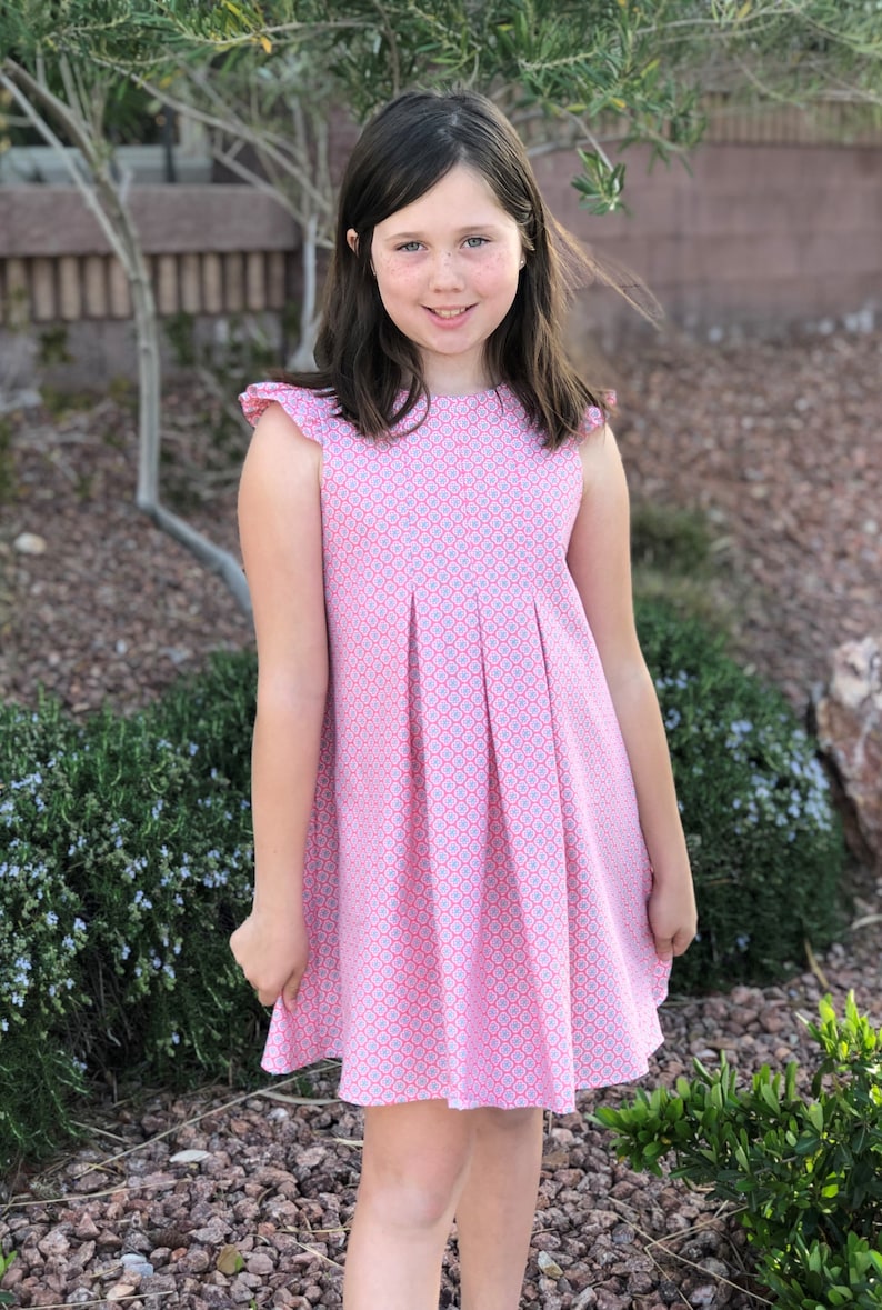 May include: A young girl wearing a pink and white patterned dress with a ruffled neckline. The dress has a pleated skirt and is sleeveless.