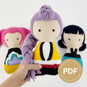 Puede incluir: Tres muñecas de fieltro hechas a mano con caras estilizadas y atuendos coloridos. La muñeca central tiene una parte superior amarilla con detalles en blanco y negro, un cinturón rojo y un peinado trenzado morado. Las otras muñecas tienen cabello y atuendos rosas y azules. La imagen incluye el texto "sewing pattern and tutorial PDF".