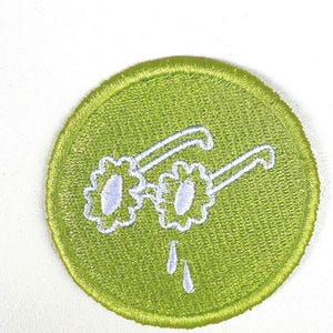 May include: A round, lime green embroidered patch featuring white sunglasses with flower-shaped lenses and two white teardrops dripping below. The patch has a stitched border and is set against a white background.