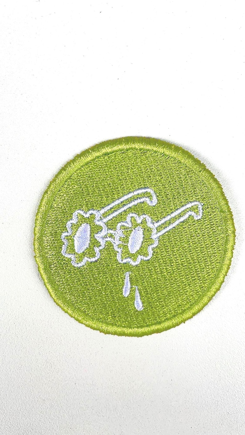 May include: A round, lime green embroidered patch featuring white sunglasses with flower-shaped lenses and two white teardrops dripping below. The patch has a stitched border and is set against a white background.