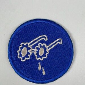 May include: Embroidered patch featuring a pair of glasses with flower-shaped lenses and teardrops, set against a blue circular background. The glasses are white, and the teardrops are white. The patch is likely for clothing or accessories.