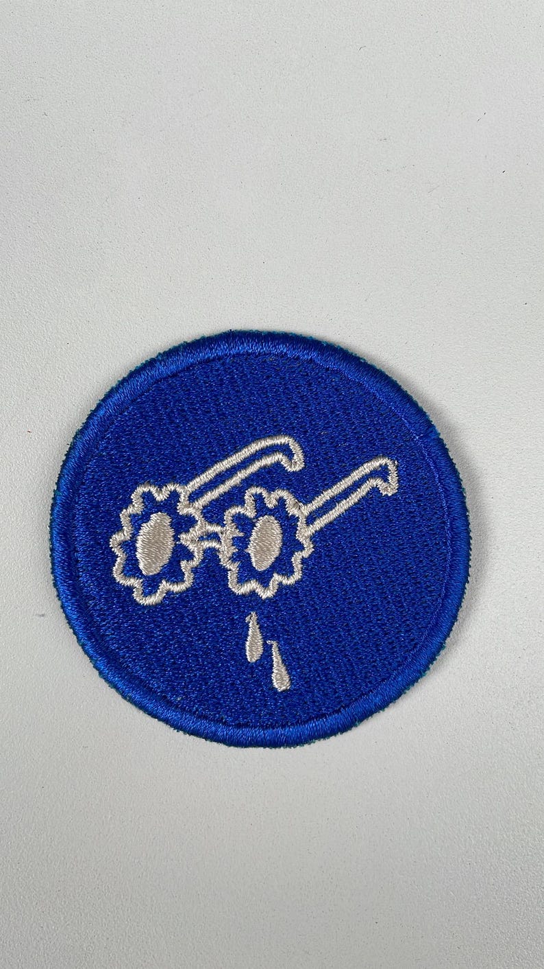 May include: Embroidered patch featuring a pair of glasses with flower-shaped lenses and teardrops, set against a blue circular background. The glasses are white, and the teardrops are white. The patch is likely for clothing or accessories.