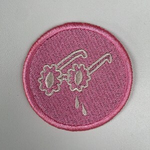 May include: A round, pink embroidered patch featuring white-outlined sunglasses with flower-shaped lenses. Two teardrops are below the glasses. The patch has a pink border and is set against a white background.
