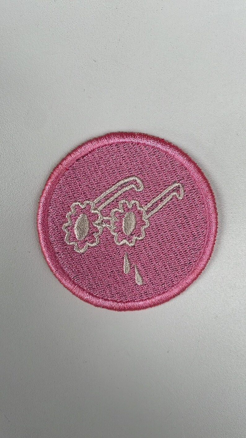 May include: A round, pink embroidered patch featuring white-outlined sunglasses with flower-shaped lenses. Two teardrops are below the glasses. The patch has a pink border and is set against a white background.