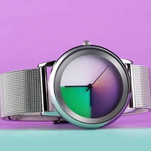 May include: A silver wristwatch with a mesh band. The watch face is divided into quadrants of white, green, and purple. The watch has a minimalist design with a single hand indicating the time.