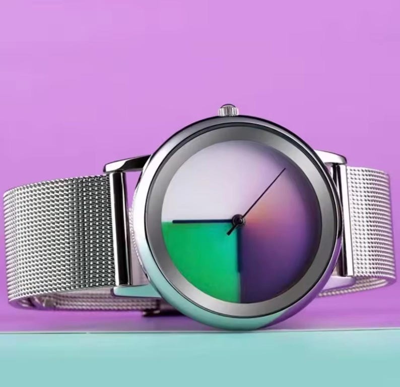 May include: A silver wristwatch with a mesh band. The watch face is divided into quadrants of white, green, and purple. The watch has a minimalist design with a single hand indicating the time.