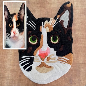 May include: A close-up of a colorful cat rug with black, white, brown, and orange fur. The rug is made of a soft, plush material and features a realistic depiction of a cat's face.