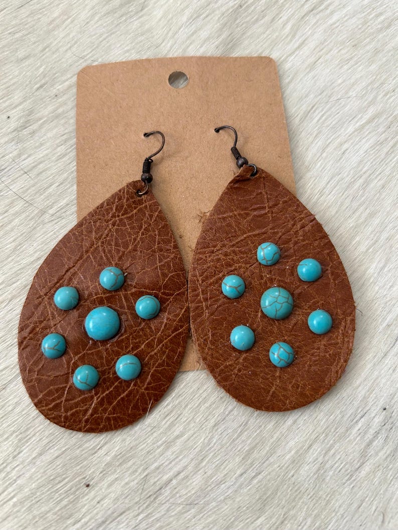 May include: A pair of brown leather earrings with turquoise stones. The earrings are teardrop shaped and have a hook closure.