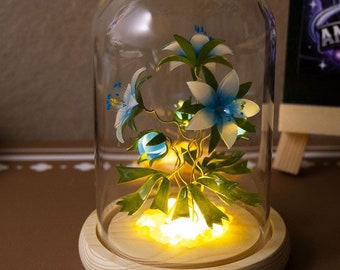 Puede incluir: Una cúpula de vidrio con una base de madera que contiene un arreglo de flores de papel con flores azules y blancas y hojas verdes. La cúpula está iluminada desde abajo con una luz blanca cálida.