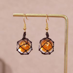 Splatoon golden egg earrings featuring handcrafted translucent resin charms that resemble the glowing fish eggs from Splatoon 3's Salmon Run mode, suspended delicately in dark brown mesh netting, and finished with gold-plated copper hook findings. These dangle earrings are designed for gamer girls and fans of Nintendo accessories who appreciate subtle geeky fashion, making them an ideal choice for casual wear, cosplay, or gift giving. Captured in soft lighting to emphasize the amber tones