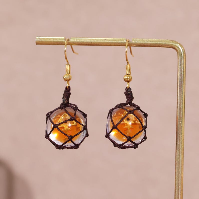Splatoon golden egg earrings featuring handcrafted translucent resin charms that resemble the glowing fish eggs from Splatoon 3's Salmon Run mode, suspended delicately in dark brown mesh netting, and finished with gold-plated copper hook findings. These dangle earrings are designed for gamer girls and fans of Nintendo accessories who appreciate subtle geeky fashion, making them an ideal choice for casual wear, cosplay, or gift giving. Captured in soft lighting to emphasize the amber tones