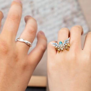 May include: Two rings are displayed. One is a silver band with small, clear stones. The other is a gold ring with a blue gemstone and wing-like accents, adorned with small, clear stones. The ring is on a finger.