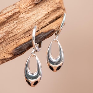 May include: A pair of silver-toned teardrop earrings. Each earring has a smooth, polished surface with a hollow, oval-shaped design. The bottom of each earring features three small, black circular accents. The earrings are displayed against a wooden background.