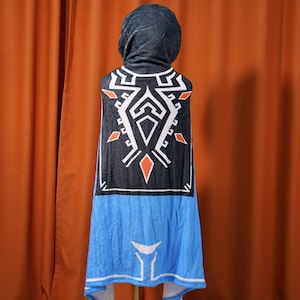 May include: A hooded fleece blanket with a blue and black design featuring a geometric pattern. The pattern includes a white diamond shape with orange and red accents.