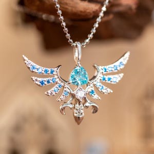 Puede incluir: Un collar con colgante plateado con un diseño de alas adornado con piedras preciosas azules y blancas. El colgante tiene una gema azul central y cuelga de una delicada cadena. Las alas están adornadas con piedras azules y blancas más pequeñas.