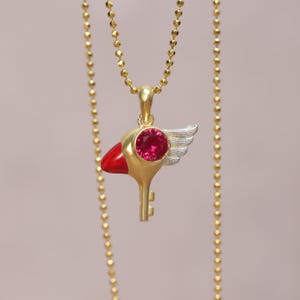 Peut inclure: Un collier en or avec un pendentif en forme de clé rouge et or orné d'une pierre précieuse rouge et d'une aile argentée.