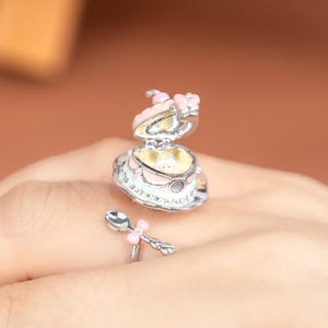 Può includere: Anello argentato con un design a forma di torta a cuore con glassa rosa e bianca e un piccolo cucchiaio con un fiocco rosa. La torta si apre per rivelare un interno giallo.