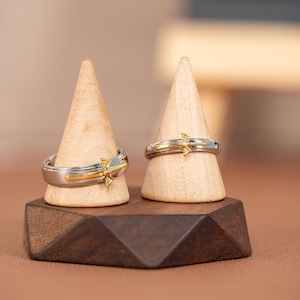 Könnte beinhalten: Zwei silber- und goldfarbene Ringe mit dekorativem Design, ausgestellt auf Holzständern. Die Ringe haben ein zweifarbiges Design mit goldenen Akzenten. Die Ringe stehen auf einem dunkelbraunen Holzsockel.