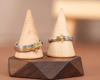 May include: Two silver and gold rings with a decorative design, displayed on wooden stands. The rings have a two-tone design with a gold accent. The rings are on a dark brown wooden base.