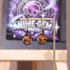 Detailed product photo showcasing the size and shape of the Splatoon golden egg earrings in comparison to a human hand, giving customers a clear idea of scale. The fish egg portion measures 1.5 inches (3.8 cm), handcrafted from high-quality resin with a soft translucent glow. The mesh netting and metal components are arranged for both durability and aesthetic appeal, ideal for Splatoon fans and cosplay accessory collectors looking for subtle game-inspired earrings.