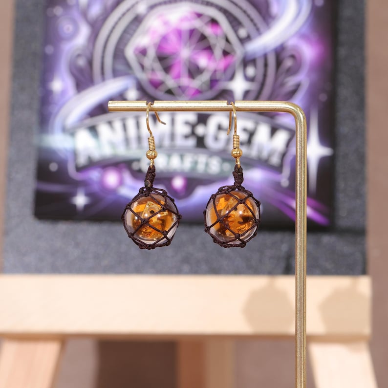 Detailed product photo showcasing the size and shape of the Splatoon golden egg earrings in comparison to a human hand, giving customers a clear idea of scale. The fish egg portion measures 1.5 inches (3.8 cm), handcrafted from high-quality resin with a soft translucent glow. The mesh netting and metal components are arranged for both durability and aesthetic appeal, ideal for Splatoon fans and cosplay accessory collectors looking for subtle game-inspired earrings.
