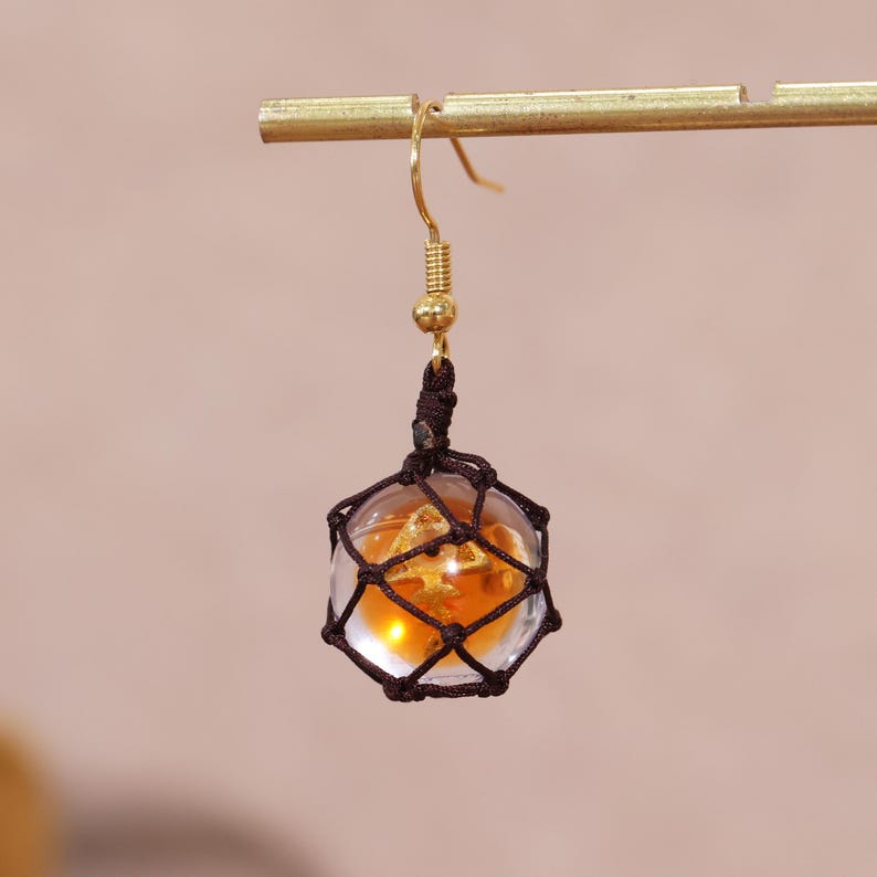 Splatoon golden egg earrings featuring handcrafted translucent resin charms that resemble the glowing fish eggs from Splatoon 3's Salmon Run mode, suspended delicately in dark brown mesh netting, and finished with gold-plated copper hook findings. These dangle earrings are designed for gamer girls and fans of Nintendo accessories who appreciate subtle geeky fashion, making them an ideal choice for casual wear, cosplay, or gift giving. Captured in soft lighting to emphasize the amber tones