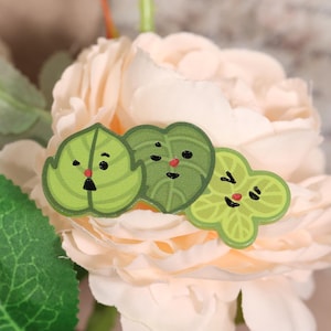 May include: Three leaf-shaped stickers with cartoon faces. The leaves are various shades of green with black eyes and mouths, and red noses. The stickers are arranged on a light pink flower, with a green leaf in the lower left corner.
