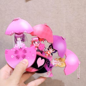 May include: A collection of pink acrylic charms featuring anime-style characters inside umbrella-shaped designs. The charms have various colors and designs, including cherry blossoms and hearts. Each charm has a small chain.