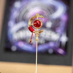 May include: A gold decorative staff featuring a red gemstone and a crescent moon design. The staff has a red tassel and a small gold bead. The background is blurred with purple and white hues.