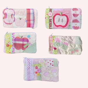 May include: Five handmade patchwork zipper pouches. Each pouch features apple or butterfly appliqués, buttons, and lace details. The colour palette includes pink, green, and white fabrics.